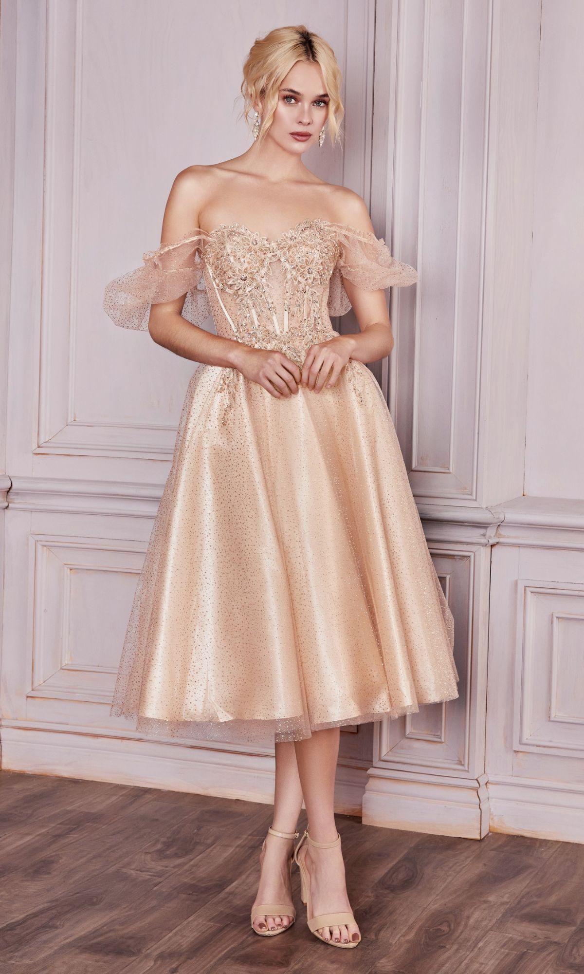 Woman wearing the Puff-Sleeve Tea-Length Prom Dress: CD0187, a shimmery blush pink midi dress with an off-the-shoulder lace bodice, stands before a light gray paneled wall on a wooden floor.